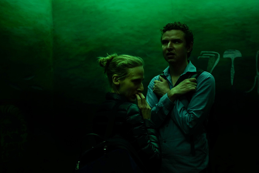 Two people pressed against a graffitied wall under a single toxic-green light: she turns toward him with a hand raised to her mouth, he grips his own crossed arms and stares past her, mouth open; everything else collapses to flat dark green