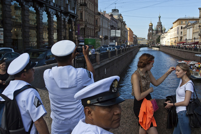 A canal embankment: three naval cadets in white uniforms crowd the left foreground, one raising a phone to photograph the water, one turned back toward the lens; at right two women stand at the parapet, one reaching up to touch the other's face; far behind, the onion domes of the Church of the Saviour on Spilled Blood