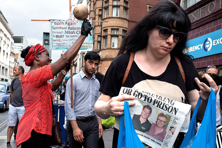 Packed London shopping street: at the bottom right a woman in cat-eye sunglasses holds up a tabloid headlined OR GO TO PRISON; a young South Asian man in a grey shirt is wedged behind her looking into the lens; behind them a woman in a red headwrap laughs head back, raising a wooden cross topped with a coconut and a Born Again placard